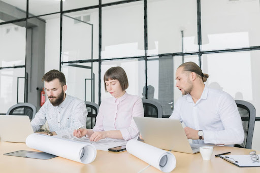 Three professionals working together at a table with laptops and rolled plans, representing teamwork, collaboration, and focused project planning in a modern office environment.