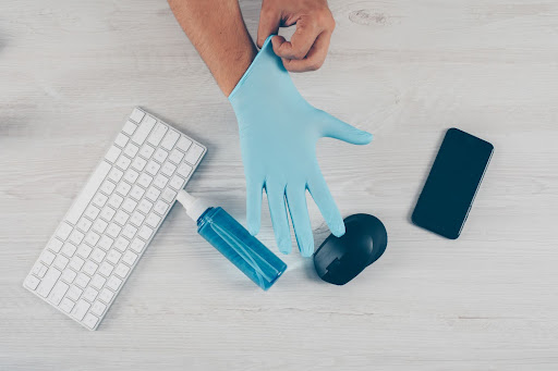 Workplace hygiene essentials on a modern desk Person putting on protective gloves beside a keyboard, hand sanitiser, smartphone, and mouse, representing hygiene and health awareness in a modern work environment.