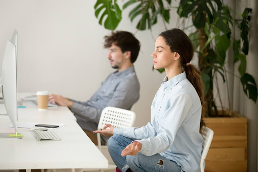 Employee practicing mindfulness meditation at a desk while a colleague works nearby, representing workplace wellbeing, recovery, and mental balance