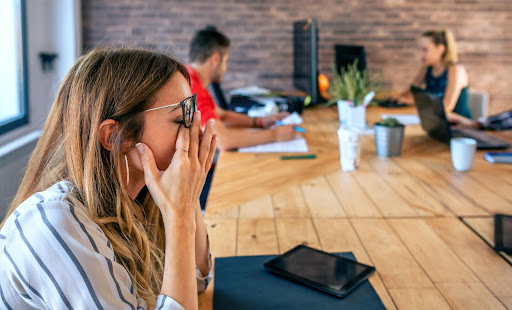 Modern workplace stress and burnout during a team meeting An employee holding her face in frustration while colleagues work in the background during a stressful office meeting