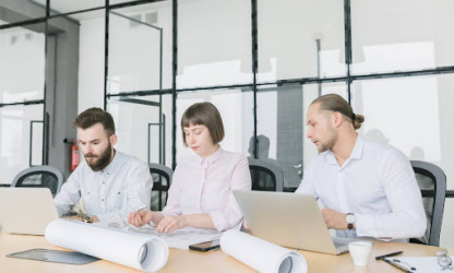 Three professionals working together at a table with laptops and rolled plans, representing teamwork, collaboration, and focused project planning in a modern office environment.