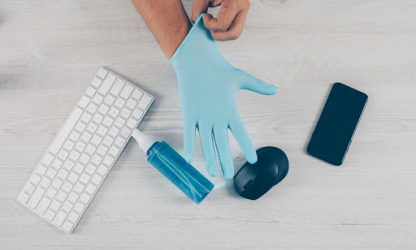 Person putting on protective gloves beside a keyboard, hand sanitiser, smartphone, and mouse, representing hygiene and health awareness in a modern work environment.