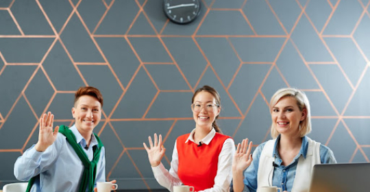Three professionals sitting at a table, smiling and waving during a meeting, representing teamwork, inclusivity, and positive workplace culture.