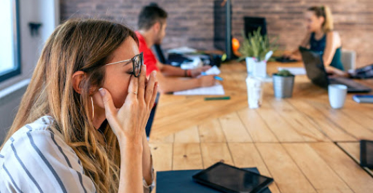 An employee holding her face in frustration while colleagues work in the background during a stressful office meeting